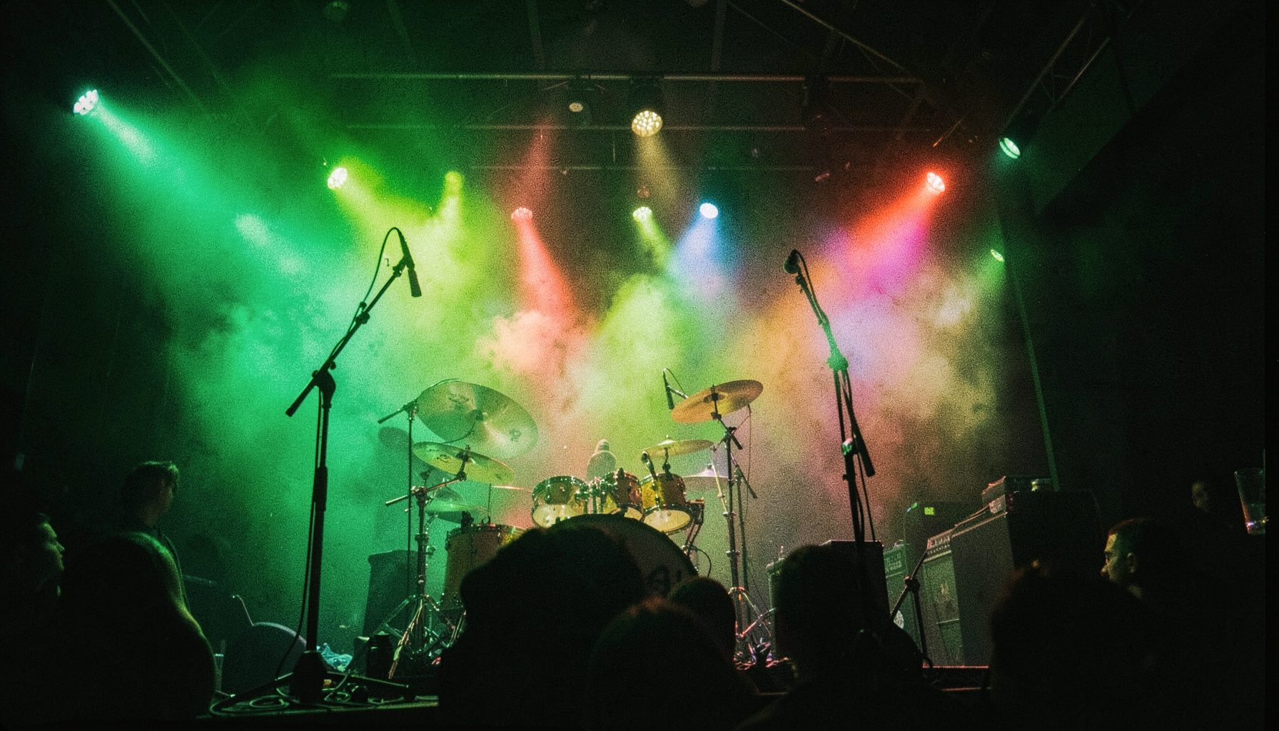 A dimly lit concert stage with colorful stage lights cutting through haze, a drum kit silhouetted in the foreground.